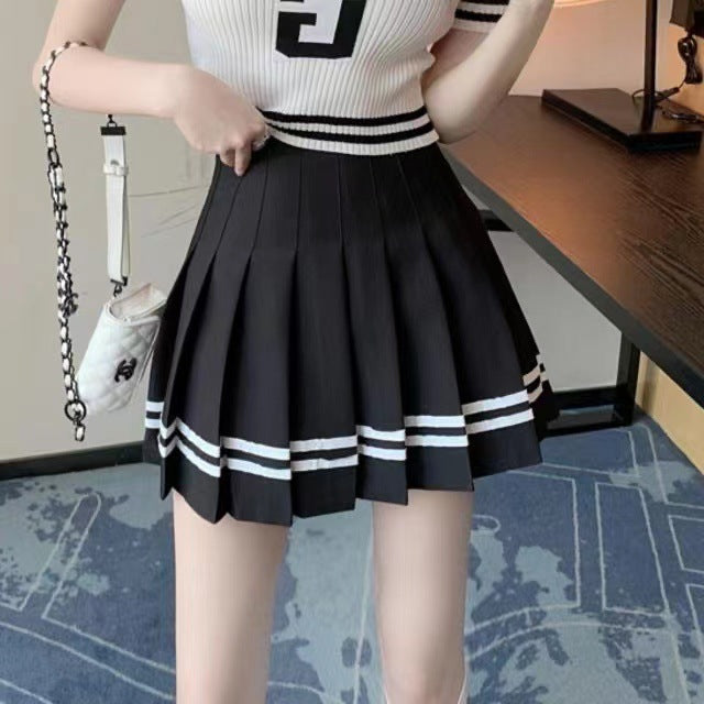 A black pleated skirt featuring white stripes near the hem, paired with a ribbed white and black top. The shot is a mid-body view, showing the skirt in a modern, casual room setting. This outfit showcases trendy femboy clothes with an athletic vibe.