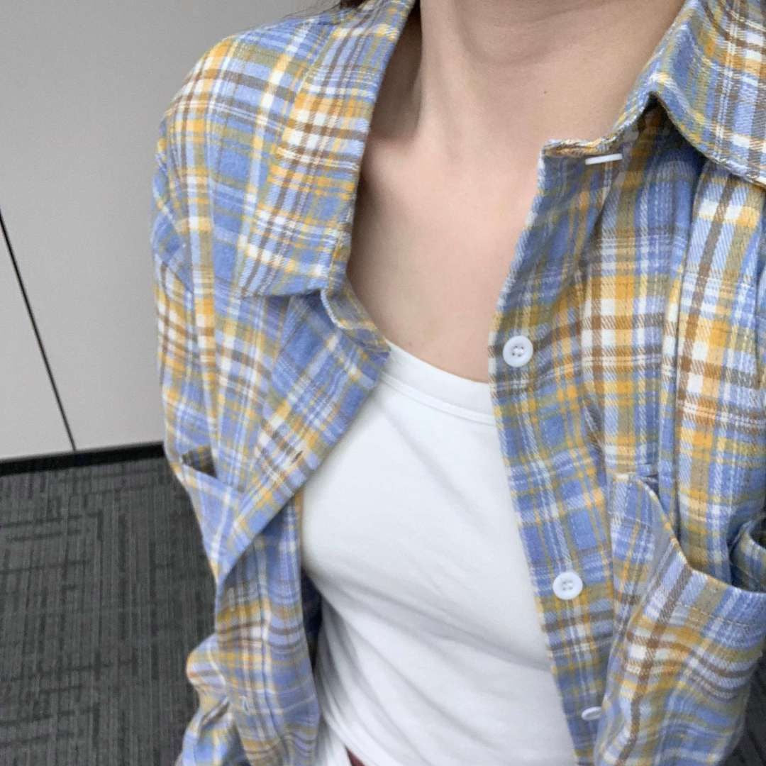 Close-up shot of a person wearing a blue and yellow plaid button-up shirt over a white tank top, showcasing the casual style. The camera angle is overhead, capturing the upper torso in natural indoor lighting. The shirt's light texture emphasizes the relaxed femboy outfit.