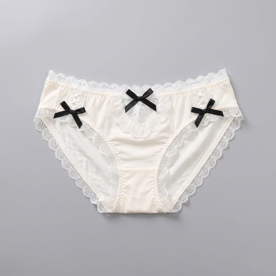 Cream lace femboy maid panties flatlay with black ribbon bow detail, scalloped lace trim