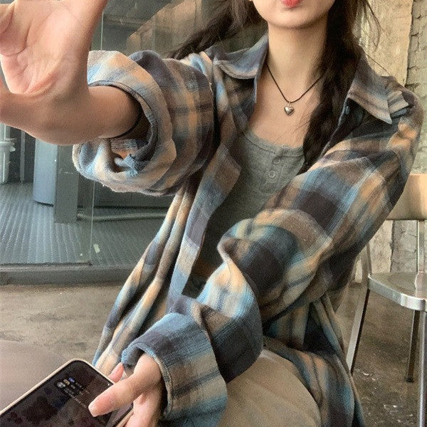A playful selfie of a person in a blue plaid button-up shirt, making a peace sign, seated in a cozy café. The image portrays the laid-back vibe of femboy fashion.