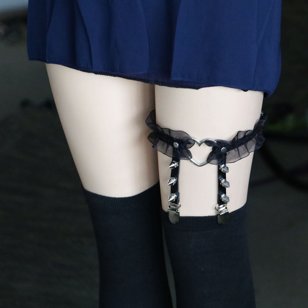 Close-up of a heart lace femboy garter worn on the thigh, featuring sheer black ruffled lace, a silver heart-shaped center ring, studded vertical straps with metal clips, and black thigh-high socks, styled in cute gothic femboy fashion and femboy accessories.