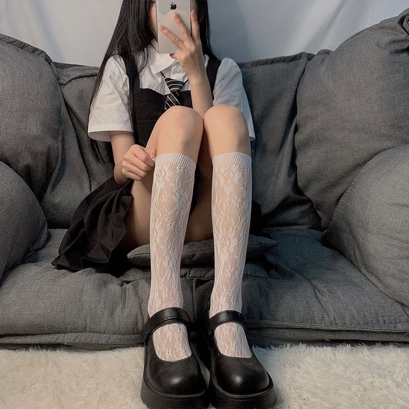 eye-level medium shot of a model seated on a gray sofa wearing white lace calf-length socks with black Mary Jane flats and a white blouse, showcasing delicate floral pattern in a chic femboy fashion look.
