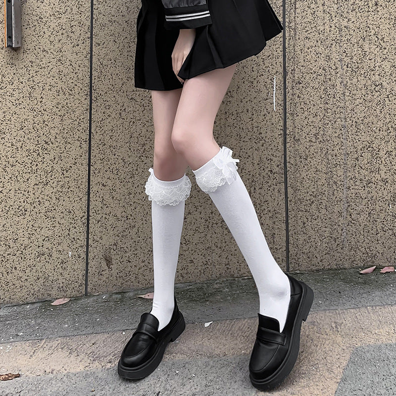 Full-length shot of black Lolita thigh-high socks with lace trim and bowknot detail, worn with black loafers. The person stands outdoors against a stone wall, presenting femboy outfits with a Lolita influence in a neutral setting.