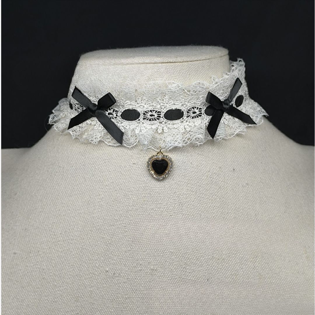 Close-up front view of a lolita lace heart choker on a mannequin, featuring white lace ruffles, black ribbon bows on each side, and a black heart-shaped pendant, styled as a cute gothic femboy accessory on a dark background.