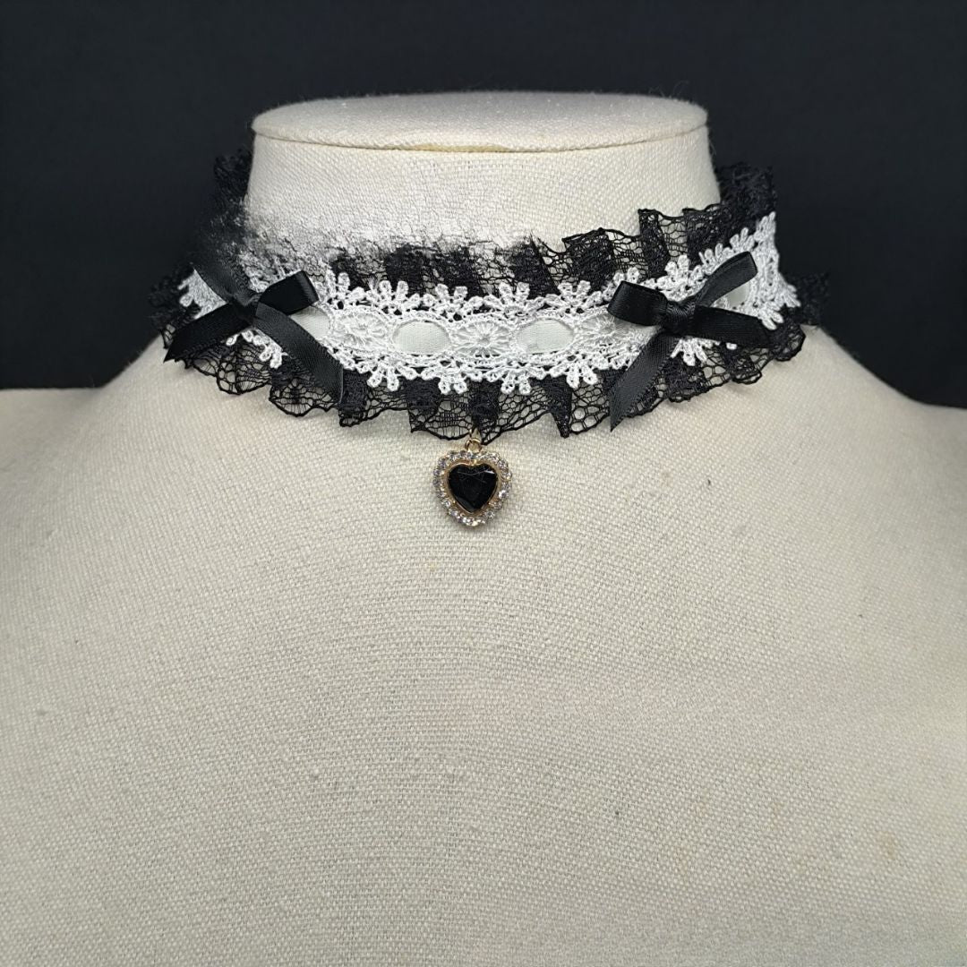Detailed close-up of a gothic lolita lace heart choker with layered black and white lace, black ribbon bows, and a black heart pendant, presented on a mannequin as a gothic femboy fashion accessory.