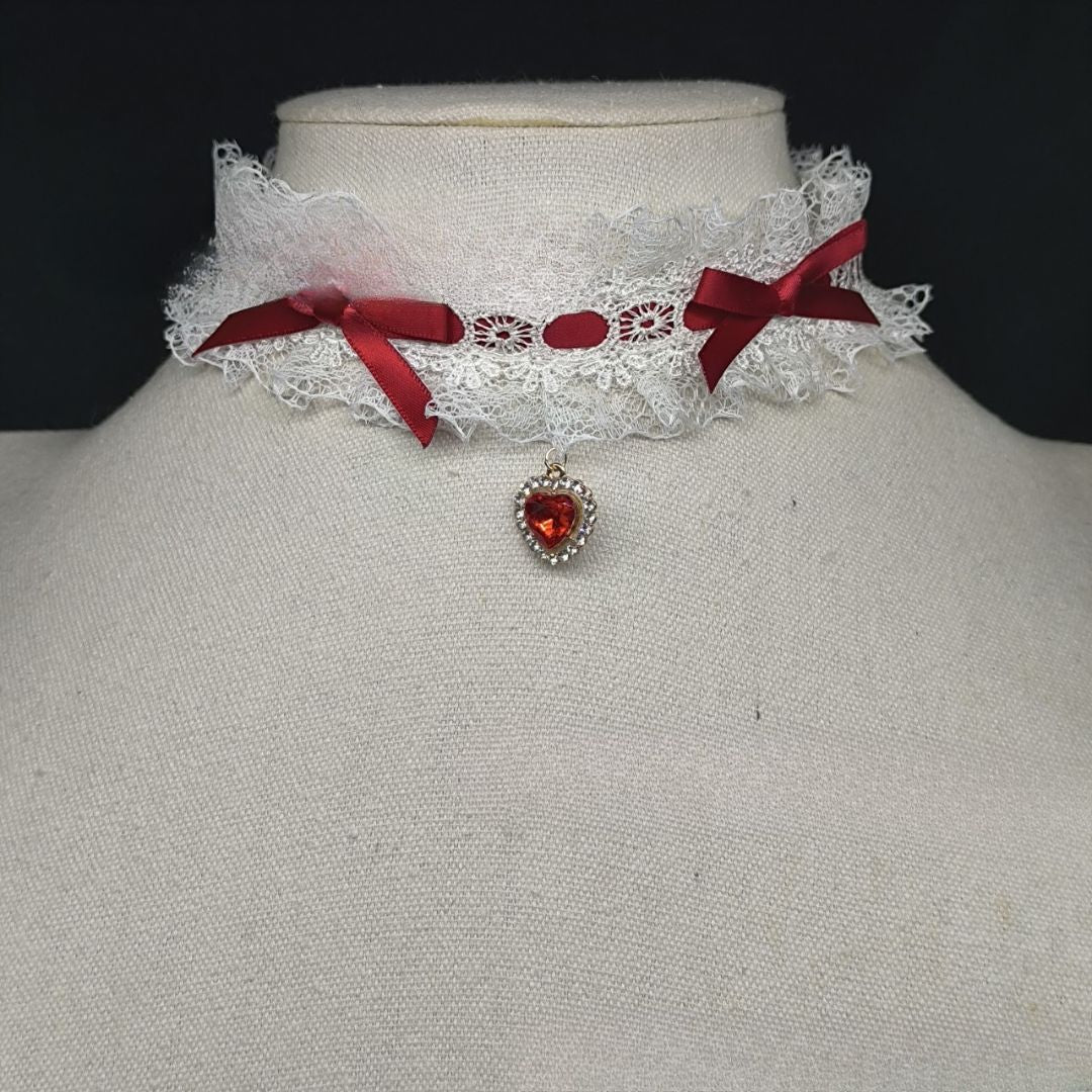 Front-facing close-up of a lolita lace heart choker featuring white lace ruffles, deep red ribbon bows, and a red heart-shaped pendant, styled on a mannequin as a romantic femboy fashion accessory.