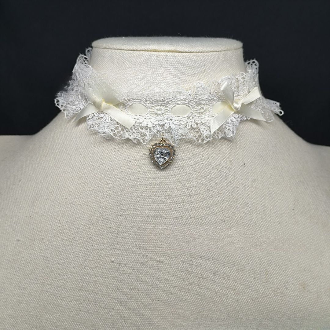 Close-up front view of a lolita lace heart choker displayed on a mannequin, designed with white lace trim, soft white ribbon bows, and a clear crystal heart pendant, creating a delicate kawaii femboy fashion accessory.