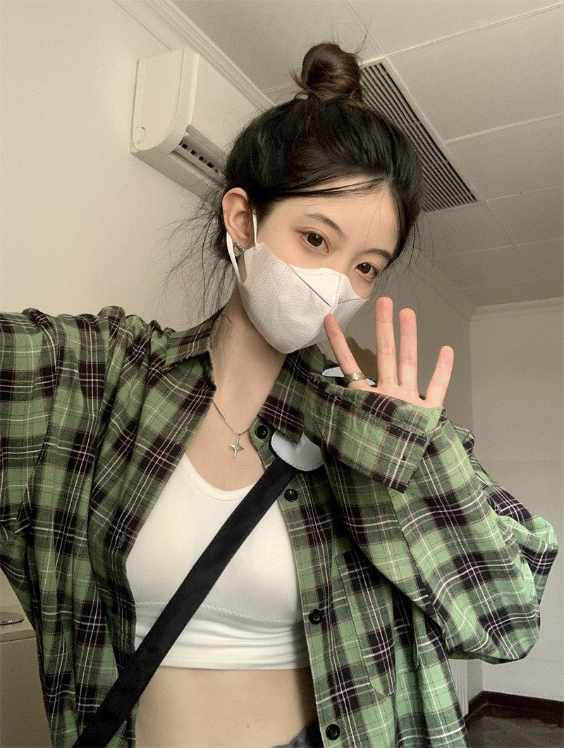 Medium shot of a model wearing a green plaid button-up shirt in femboy style, paired with a white crop top. The model is posing indoors with a casual, laid-back vibe, showcasing femboy clothing.