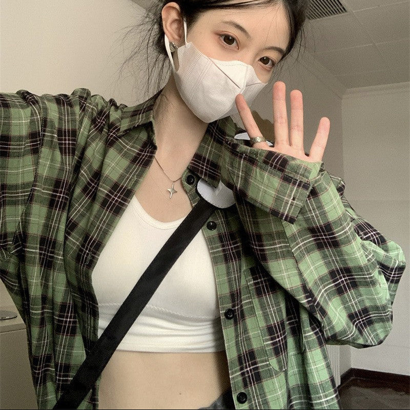 Medium shot of a model wearing a green plaid button-up shirt in femboy style, paired with a white crop top. The model is posing indoors with a casual, laid-back vibe, showcasing femboy clothing.