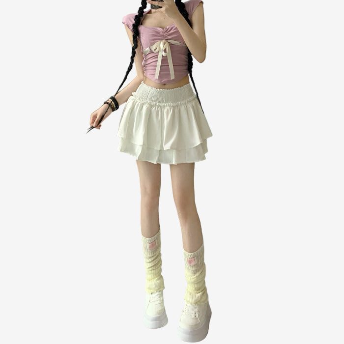 Close-up of a model wearing a white ruffle skirt, paired with a pink crop top and socks, set in a modern indoor living room. The skirt's layered design enhances a playful, feminine style, perfect for femboy outfits and femboy fashion enthusiasts seeking comfortable yet trendy femboy clothing.