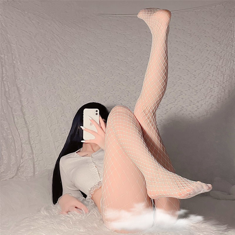 Side-angle kneeling pose of a model wearing white wide-mesh fishnet stockings paired with a pink plaid mini skirt; soft indoor lighting enhances the feminine femboy outfits and femboy clothing style.