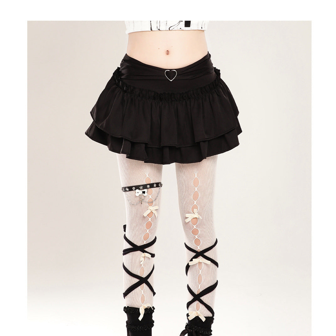 A model's lower body wearing a black ruffled mini skirt, white lace-up tights with black criss-cross straps, black socks, and black platform shoes against a light gray studio background.