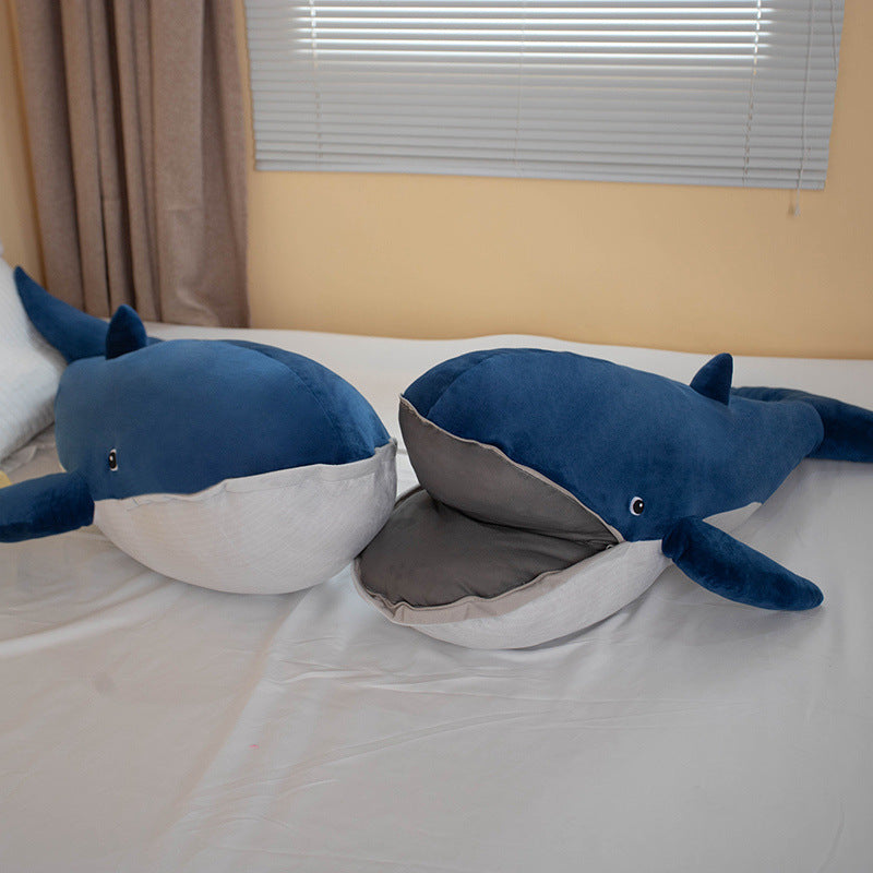 Wide bedroom lifestyle shot of two Femboy BLÅVINGAD whale plush toys resting on a bed, photographed from a slightly elevated angle with warm lighting and neutral linens, creating a relaxed, cute aesthetic commonly associated with femboy apparel branding.
