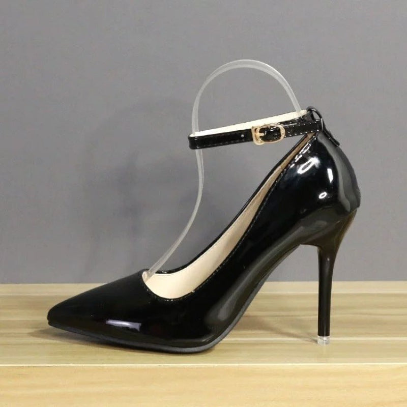 Detailed image of shiny black leather heels with a 10 cm pointed heel and sleek ankle strap, presented on a neutral grey background. Versatile choice for any femboy wardrobe.