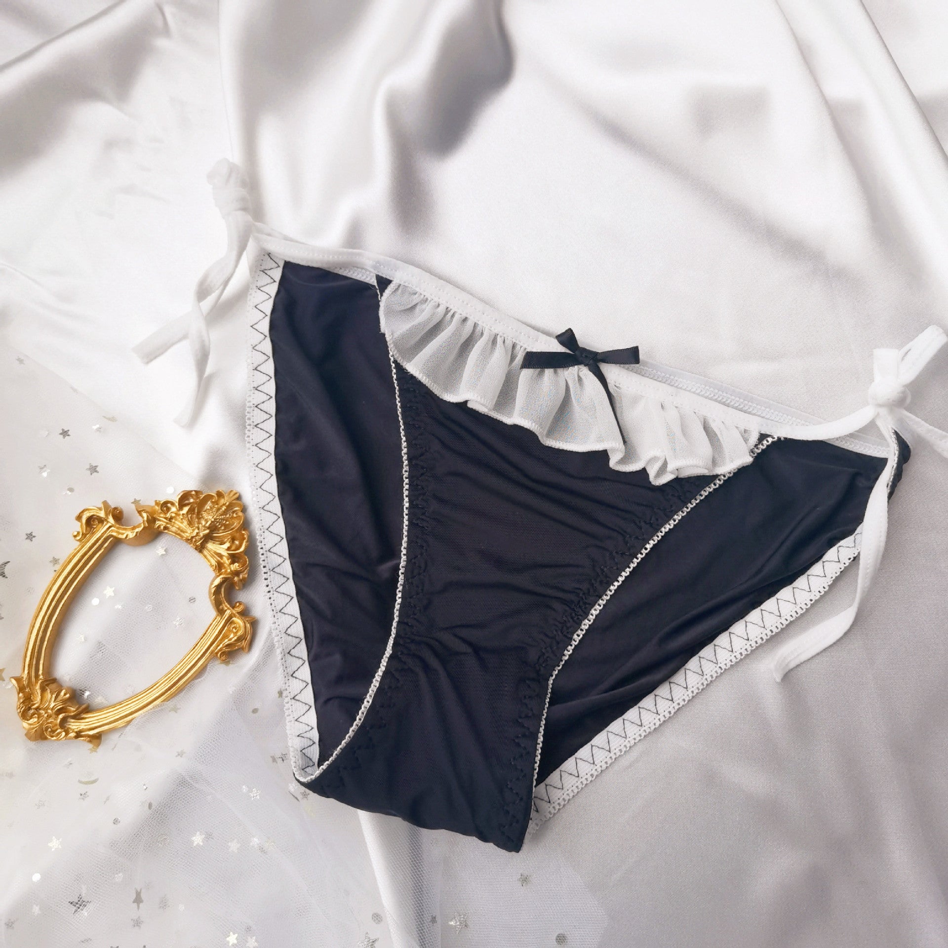 Flat lay of black Lolita maid panties with white lace ruffle trim and black bow on satin fabric – sexy femboy lingerie, best femboy panties