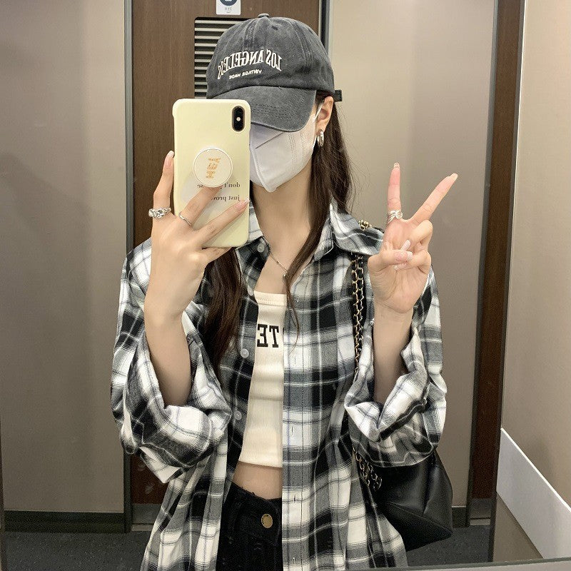 Medium shot from a mirror selfie angle showing a person wearing a black-and-white plaid button-up shirt, paired with a cap and mask, adding a street-style edge to the femboy outfit. The plaid pattern pops against the minimalist background.