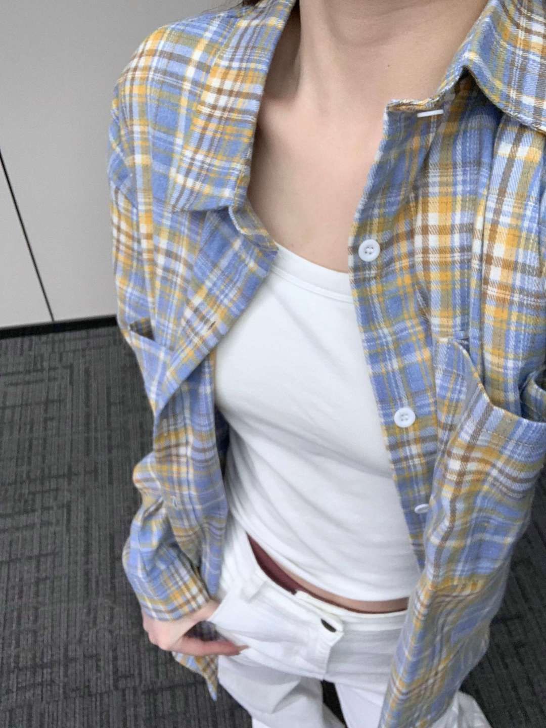 Close-up shot of a person wearing a blue and yellow plaid button-up shirt over a white tank top, showcasing the casual style. The camera angle is overhead, capturing the upper torso in natural indoor lighting. The shirt's light texture emphasizes the relaxed femboy outfit.