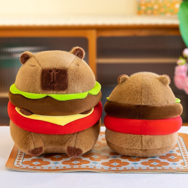 Front and back view of the Capybara Burger Plush showing a cute capybara plush designed as a hamburger with lettuce, cheese, and tomato layers, photographed on a patterned mat in a cozy indoor setting.