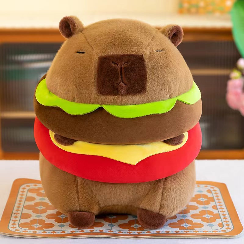 Front-facing hero image of the Capybara Burger Plush featuring a cute capybara plush styled as a hamburger with lettuce, cheese, and tomato layers, photographed on a decorative mat.
