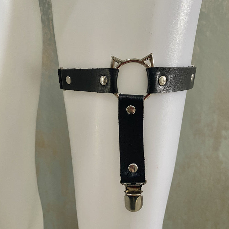 Front view of a cat face femboy thigh garter worn on a white mannequin leg, featuring black faux leather straps with silver rivet studs, a metal cat-ear ring detail at the center, and an adjustable garter clip, showcasing cute gothic femboy fashion and femboy accessories.