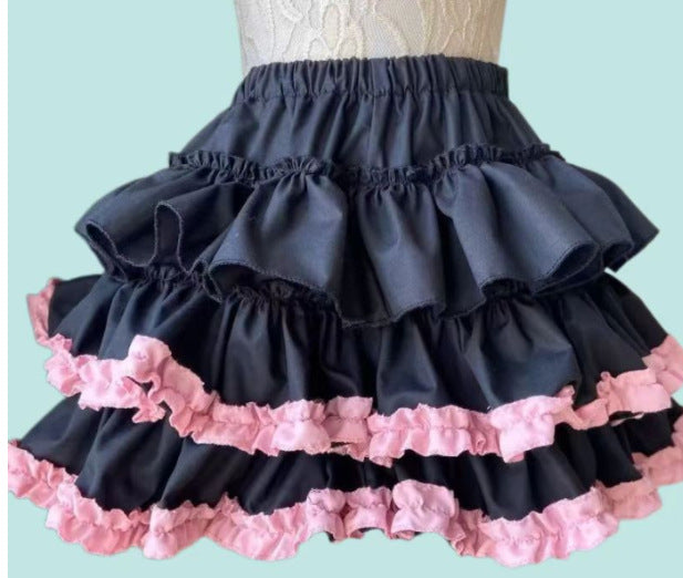 Front view of a dark punk tiered femboy skirt with layered black ruffles and soft pink frill trim, displayed on a mannequin against a pastel blue background, showcasing edgy femboy fashion styling.