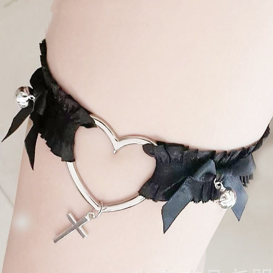 Close-up of the black Femboy Cross and Heart Garter wrapped around the thigh, featuring a heart-shaped metal design with a dangling cross. The delicate black ribbon bows and silver bell details emphasize the gothic and cute style, perfect for femboy fashion.