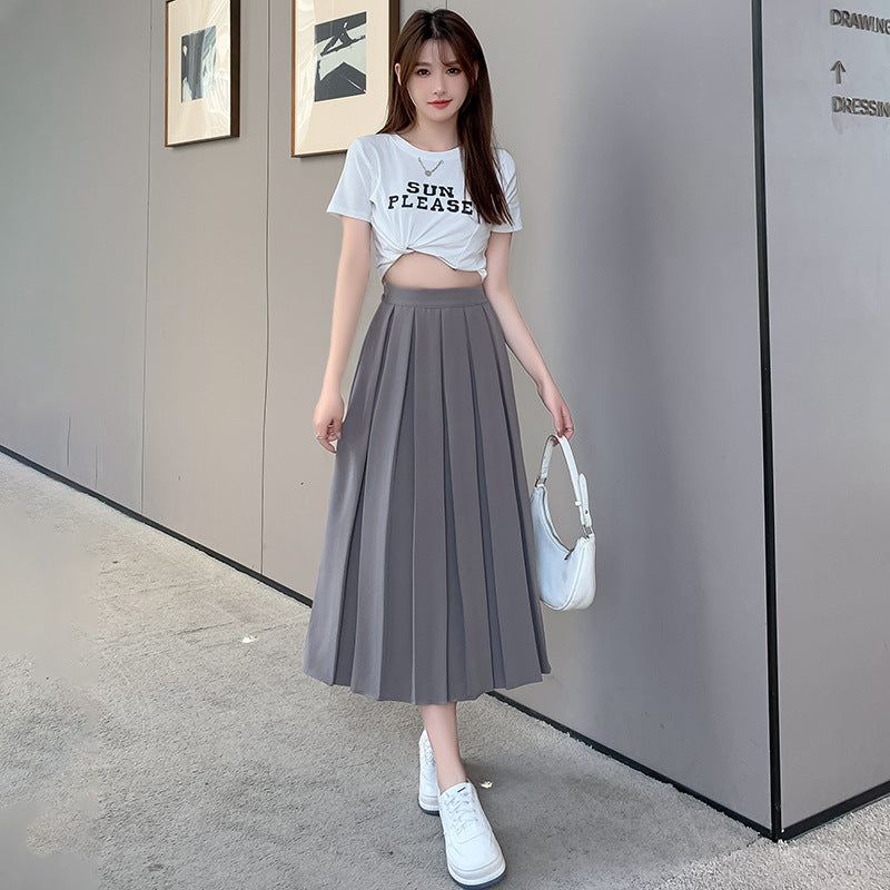 Full-body shot of model wearing a high-waisted gray A-line pleated skirt, paired with a casual white cropped tee in a modern minimalist setting. Ideal femboy clothing for a chic, casual look.