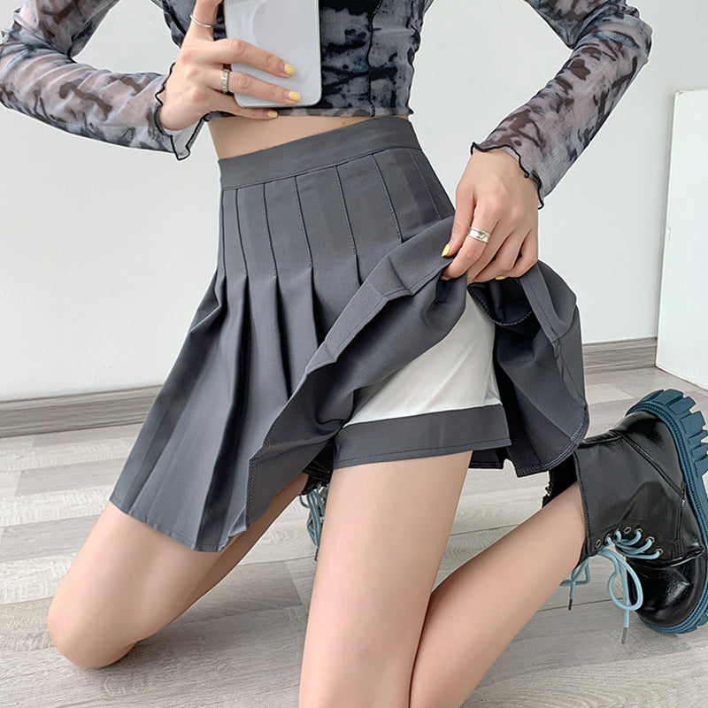 Close-up shot of a model wearing a gray high-waist pleated skirt, paired with a crop top. The model kneels on the floor and lifts part of the skirt, revealing a pair of white undershorts underneath. The image highlights the pleated design, ideal for femboy clothing and femboy outfits. The clean, minimalist background accentuates the stylish nature of this skirt, making it a standout piece in femboy fashion.