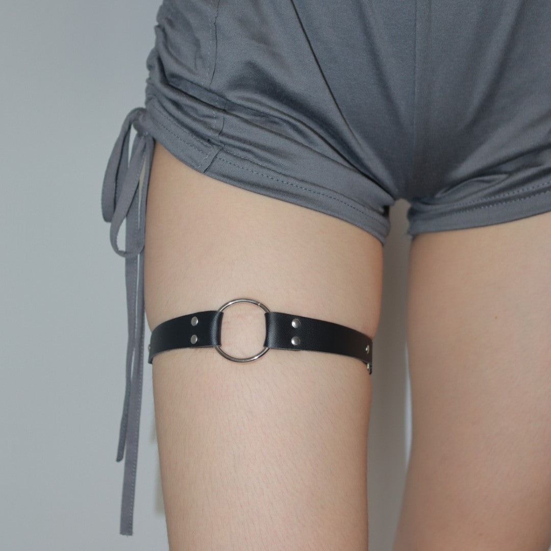 Close-up view of a femboy thigh wearing a black leather ring garter against a gray background. The femboy garter is paired with loose gray shorts, styled for femboy fashion.