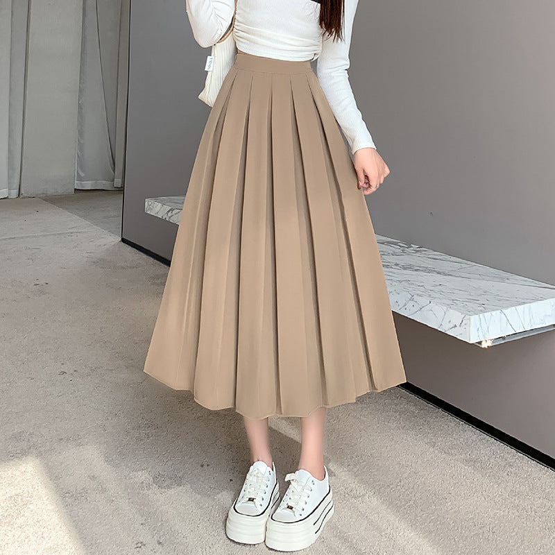 Mid-length shot of model wearing a beige high-waisted pleated A-line skirt with a fitted white top, in a modern and softly lit indoor space. Great choice for femboy fashion, combining elegance and simplicity.