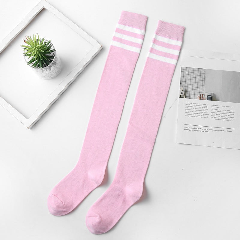 Flat-lay shot of pink thigh-high socks with white stripes at the top, styled on a white background alongside a magazine and a potted plant. This close-up captures the softness and cute aesthetic, perfect for femboy clothing styles. The arrangement highlights the delicate and playful side of femboy fashion.