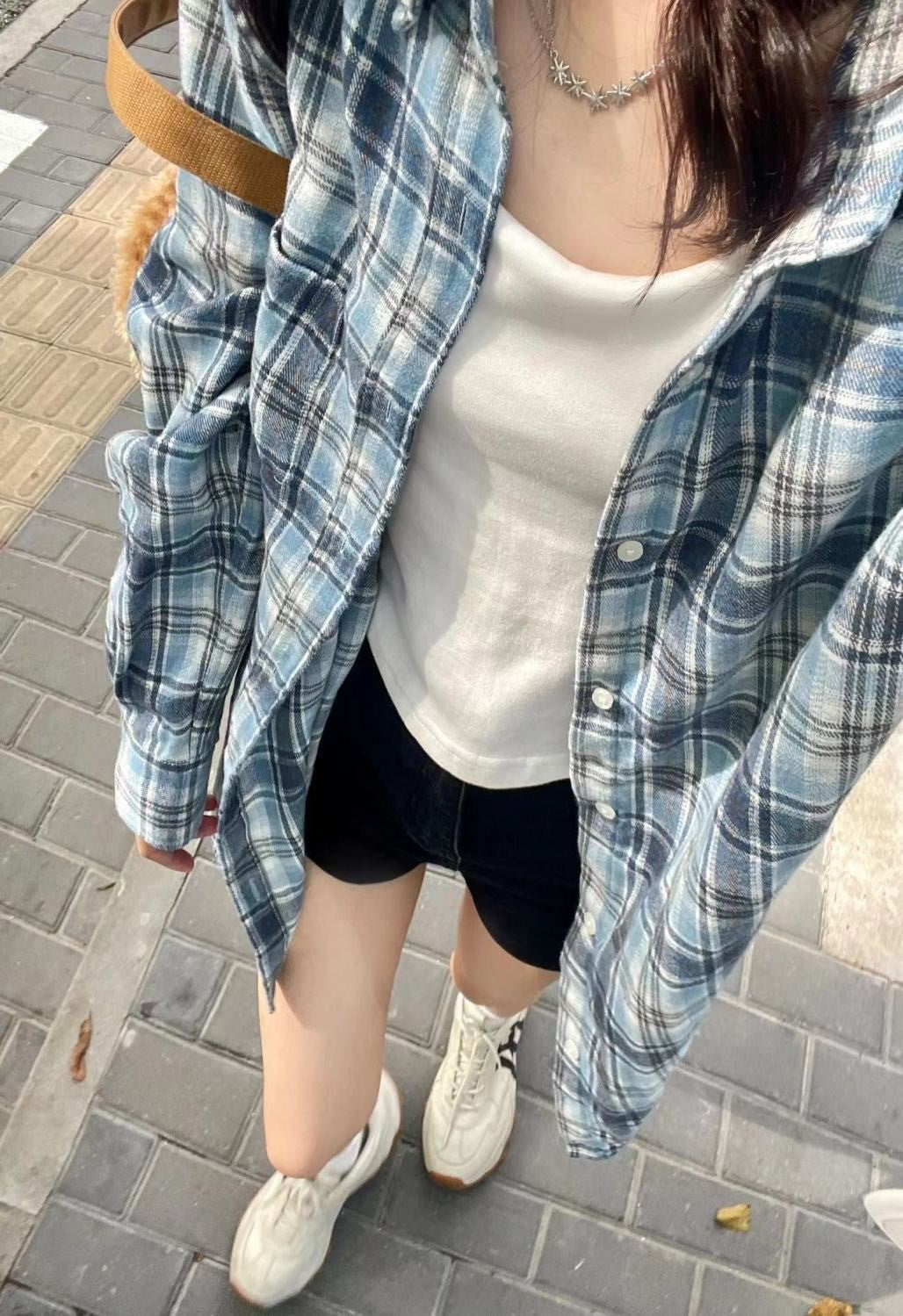 Overhead view of a person wearing a blue plaid button-up shirt over a white tank top, styled with black shorts and sneakers, walking on a city street. The shot highlights the versatility of femboy outfits.