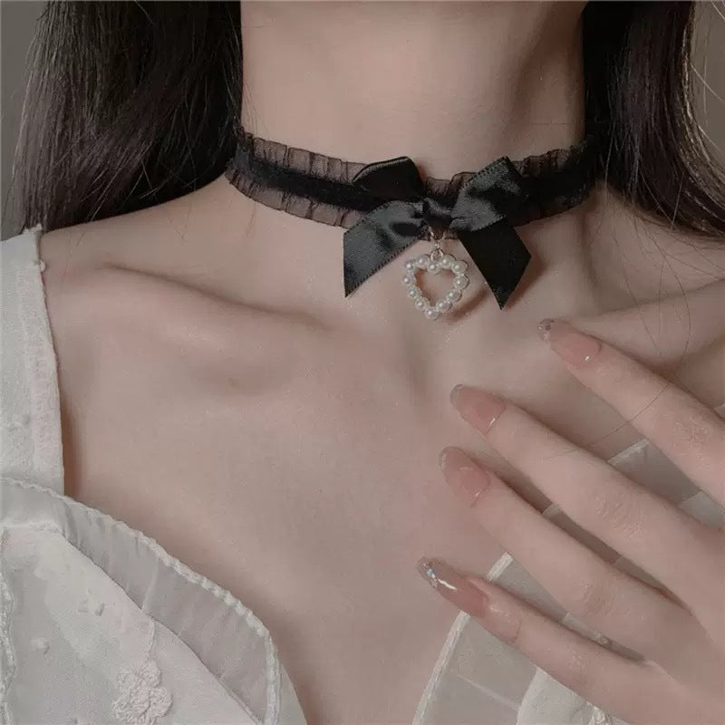 Close-up front view of a black gothic bow lace femboy choker worn on a model’s neck, featuring sheer black lace trim, a satin bow, and a pearl heart pendant, styled for gothic femboy fashion with a soft yet dark aesthetic.