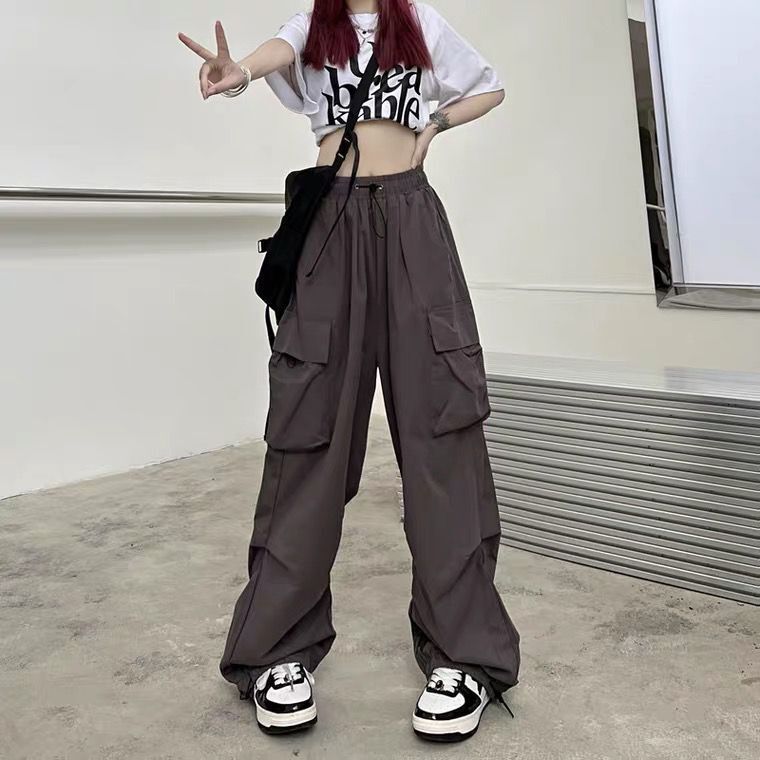 Full-body shot of model wearing dark grey parachute-style cargo pants with a drawstring waist, styled with a cropped graphic T-shirt and sneakers, set in an industrial-style interior – femboy clothes, edgy streetwear, unisex fashion trends.