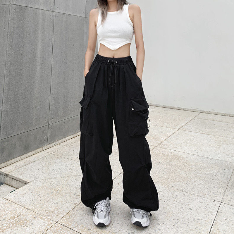 Full-body shot of model in black cargo pants with multiple pockets and drawstrings, styled with a white crop tank top, standing against a minimalist grey wall – femboy outfits, unisex streetwear, stylish cargo pants.