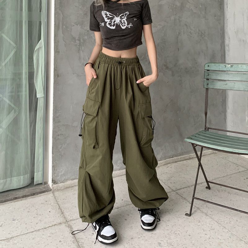 Front view of model wearing high-waisted olive green cargo pants with oversized pockets, paired with a butterfly print crop top, photographed in natural light against a grey concrete wall – femboy clothing, femboy outfits, gender-neutral fashion.