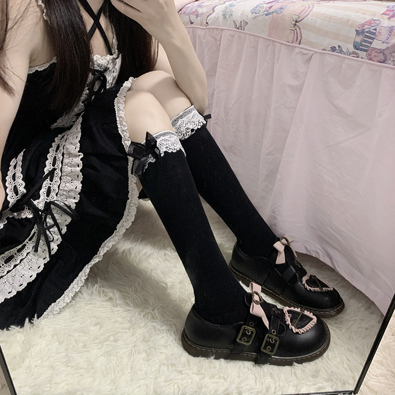 Medium shot of black Lolita thigh-high socks with lace and bow trim, matched with black gothic-inspired shoes. The person sits on a white fluffy carpet, embracing gothic Lolita vibes in femboy clothing within a pastel-themed room.