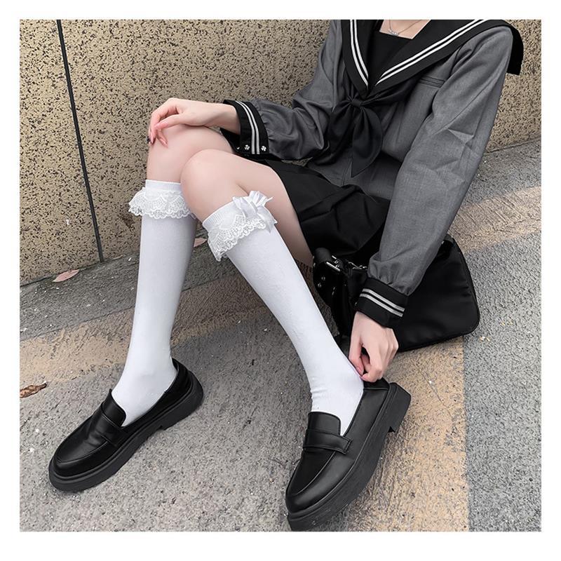 Close-up shot of white thigh-high Lolita socks with lace trim and bowknot detail, paired with black shoes. Set against a stone background with the person sitting on the ground, showcasing femboy style in a classic outdoor setting.