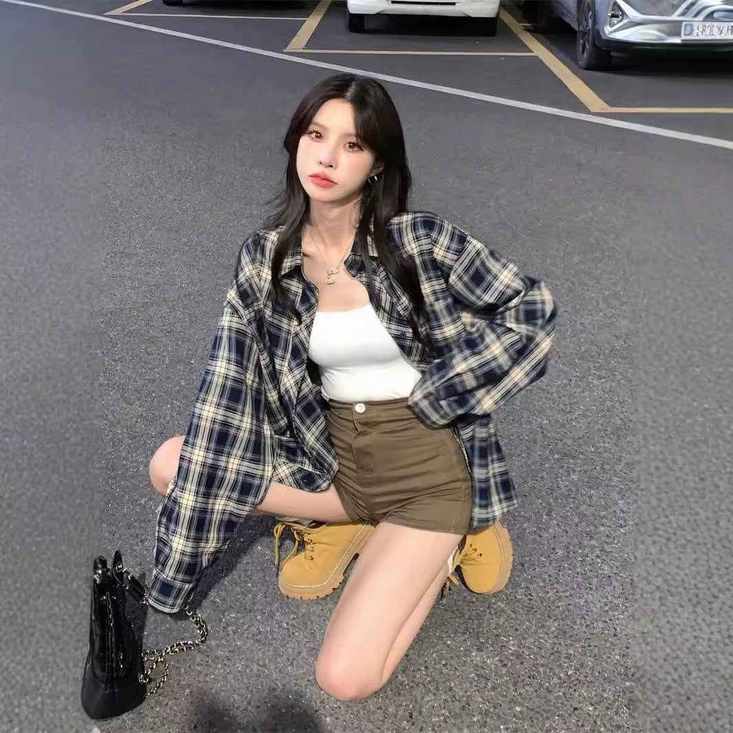 Full-body shot of a model kneeling outdoors while wearing a blue plaid button-up shirt paired with a white top and brown shorts. The image highlights a casual femboy fashion look suitable for streetwear outfits.