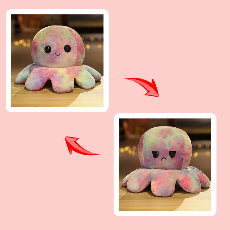 Front-facing lifestyle product shot of a pastel tie-dye reversible octopus plushie with a happy expression, photographed at eye level on a wooden surface with warm bokeh lighting, featuring soft pink, blue, and yellow tones, a cute décor accessory from a femboy clothing store.