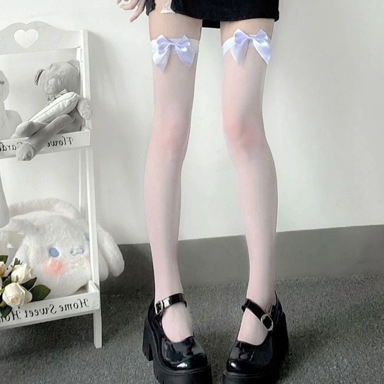 A full-body shot of a person standing and wearing white sheer thigh-high stockings with large satin bowknots near the top, paired with black platform shoes. The background features a minimal, indoor setup with plush toys and a decorative white shelf, giving a soft, cute aesthetic. Keywords: femboy clothing, femboy outfits, femboy clothes.