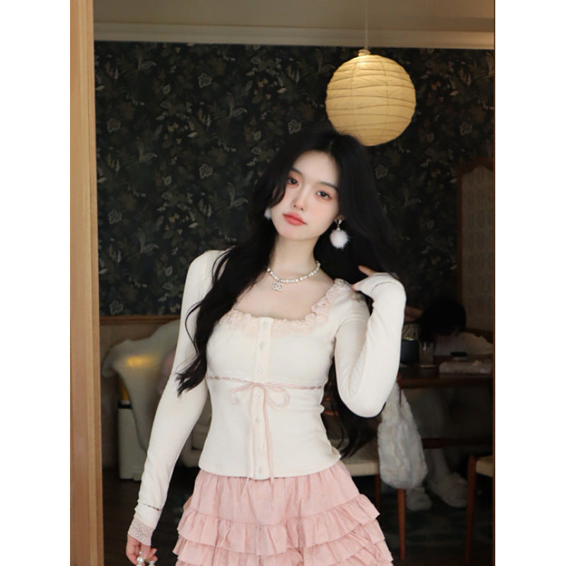 Close-up shot of a white Lace Cardigan Top with delicate lace accents, button details, and a ribbon tie at the waist, styled with a layered pink skirt. Captured indoors against a floral wallpaper background, showcasing femboy clothing and femboy fashion.