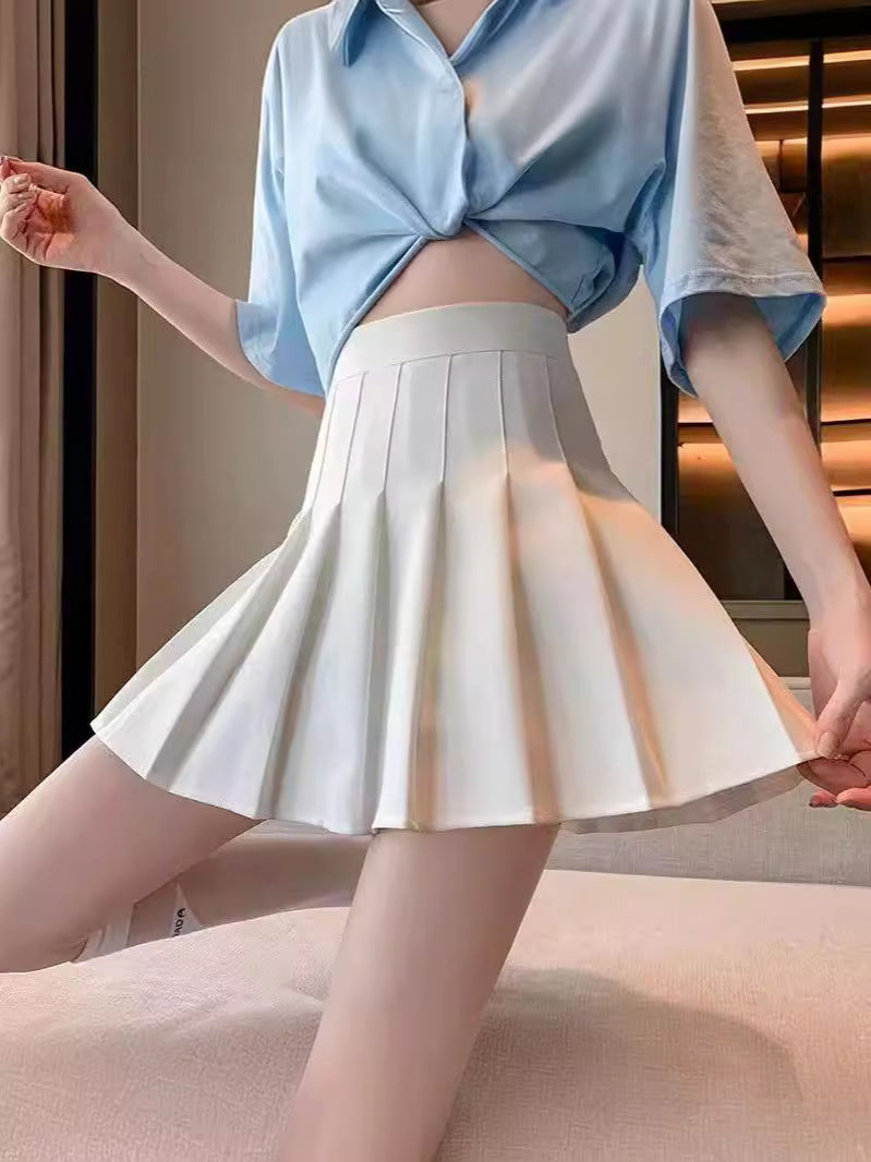 A white pleated skirt worn by a model paired with a light blue cropped shirt, set against a neutral, warmly lit indoor background. The clean pleats of the skirt add a sophisticated touch, making it a perfect piece for elegant femboy outfits and fashionable femboy clothing.